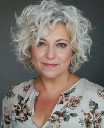 me with short curly white hair wearing a floral patterned top is looking at the camera against a dark background.